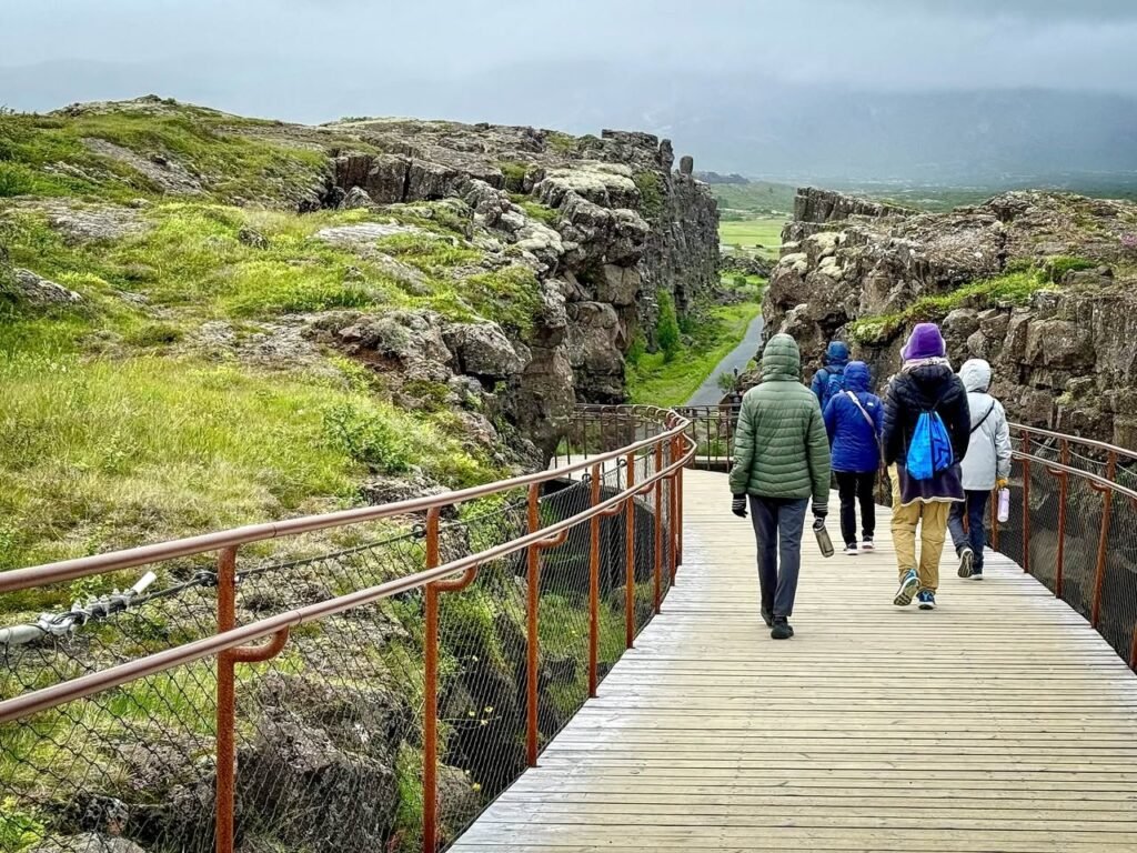 A wooden boardwalk leads through a rugged rock canyon with hikers walking towards the distant landscape under a cloudy sky.