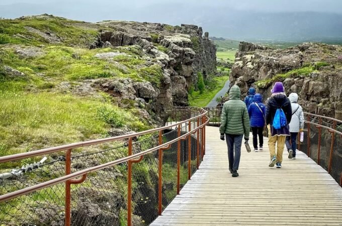 A wooden boardwalk leads through a rugged rock canyon with hikers walking towards the distant landscape under a cloudy sky.