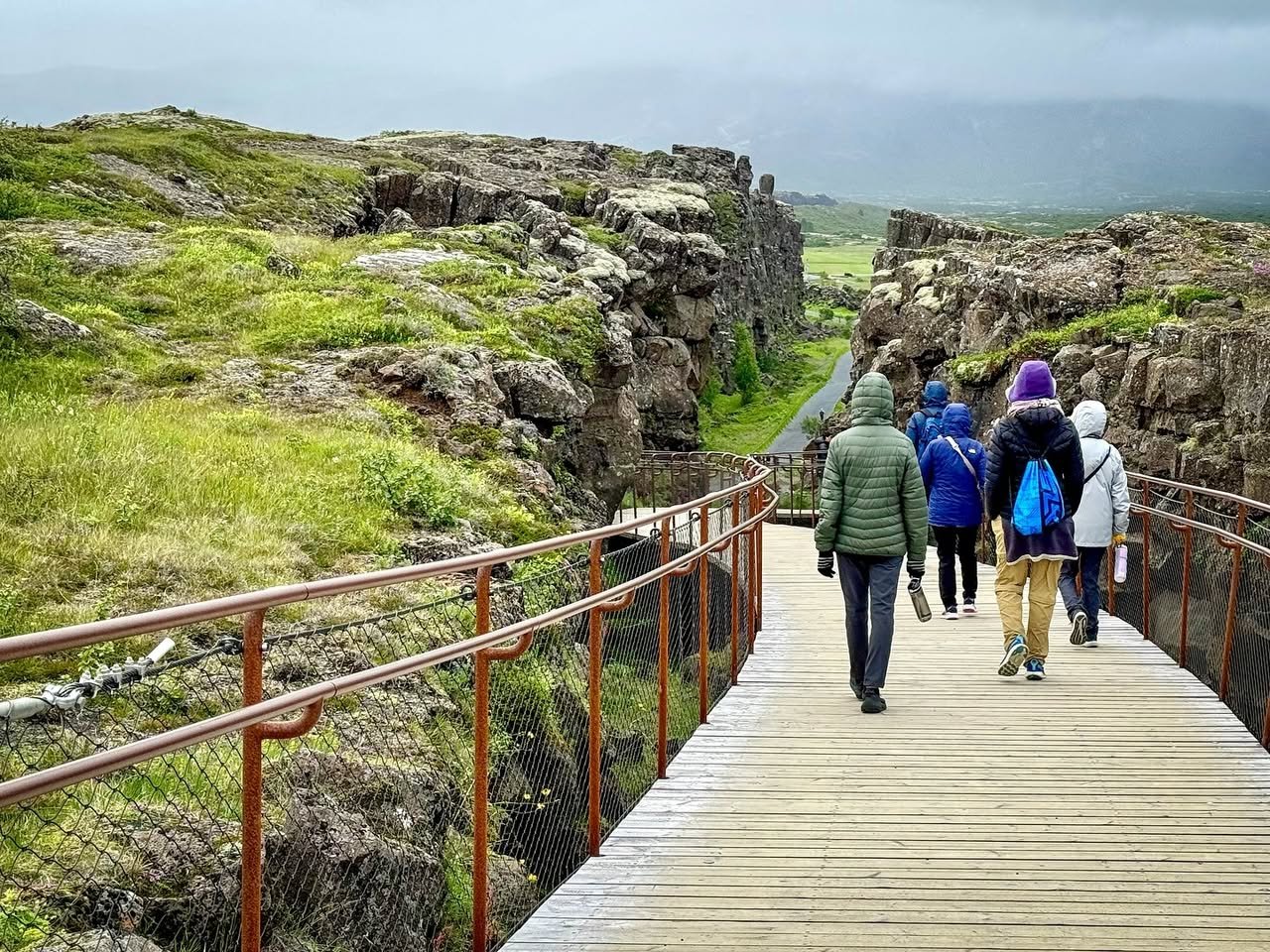 Þingvellir A wooden boardwalk leads through a rugged rock canyon with hikers walking towards the distant landscape under a cloudy sky.