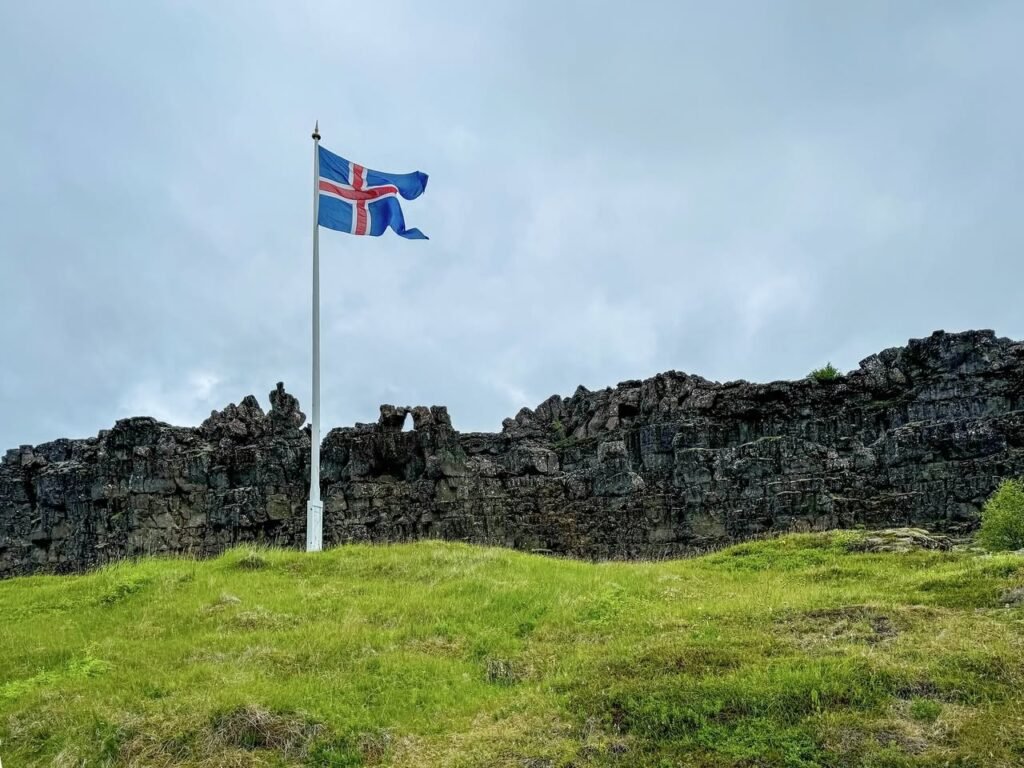 An Icelandic flag flies proudly beside an ancient drystone wall and green grass field.