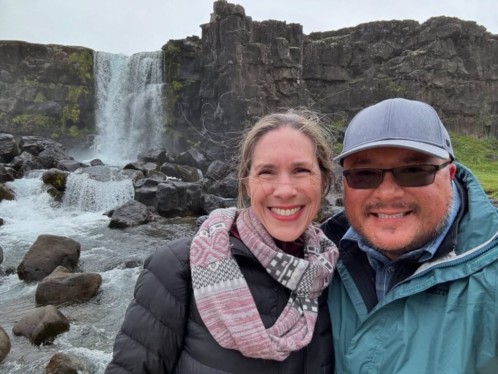 A couple takes a snapshot in front of a rugged waterfall cascading over rocky cliffs.