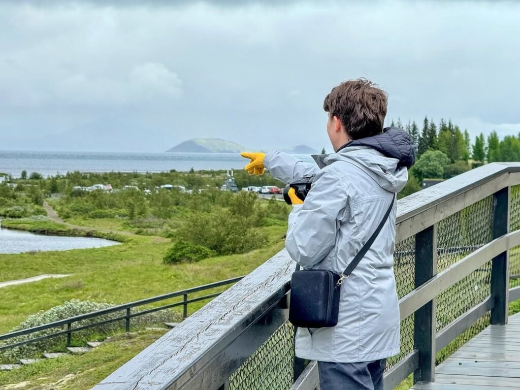 A person standing on a wooden boardwalk points towards a distant island across a body of water in a coastal wetland landscape.