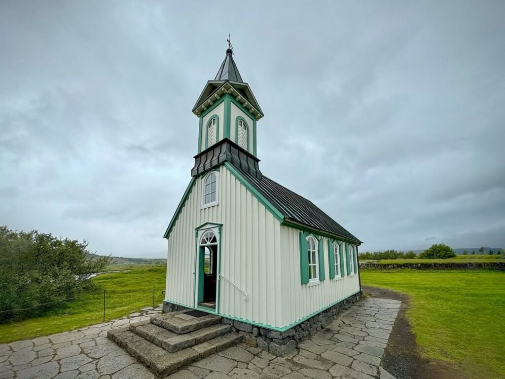 A white and green church building with a tall steeple stands on a stone path in a lush green field under an overcast sky.