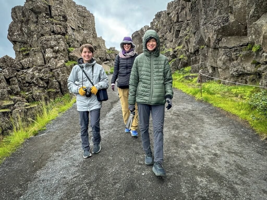 A group of three people walks along a gravel path surrounded by towering rock formations and green grass. They are dressed in winter jackets and hoods, ready for a day of outdoor exploration in a rugged landscape.
