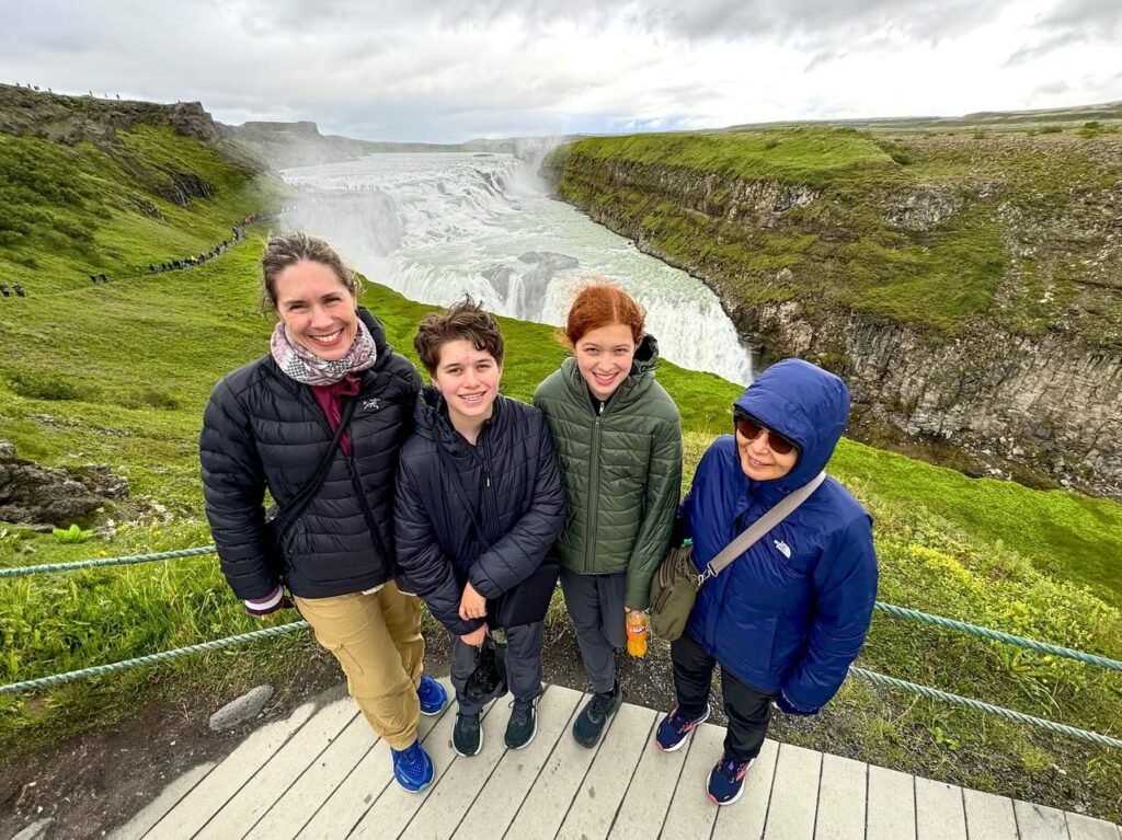 A group of travelers poses on a wooden viewing platform in front of a powerful Icelandic waterfall surrounded by lush green hills.