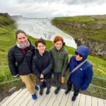 Þingvellir A group of travelers poses on a wooden viewing platform in front of a powerful Icelandic waterfall surrounded by lush green hills.