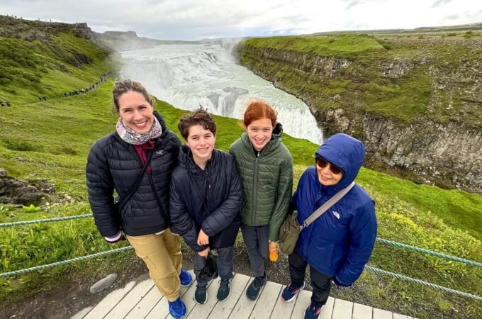 A group of travelers poses on a wooden viewing platform in front of a powerful Icelandic waterfall surrounded by lush green hills.