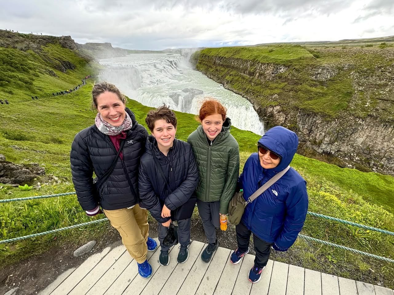 Gullfoss Waterfall A group of travelers poses on a wooden viewing platform in front of a powerful Icelandic waterfall surrounded by lush green hills.