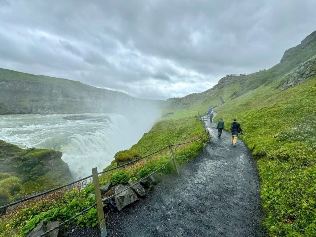Hikers walk on a gravel path beside the powerful Golfan Falls in Iceland's Snæfellsnes peninsula during a rainstorm. The misty waterfall and green highlands create an atmospheric landscape.