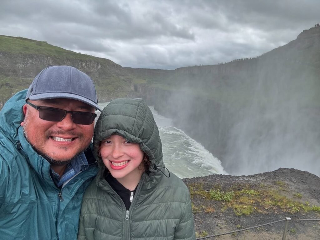 A smiling traveler stands by a misty waterfall in a windswept mountain landscape, capturing a shared adventure photo from a Pacific Northwest or international natural wonder.