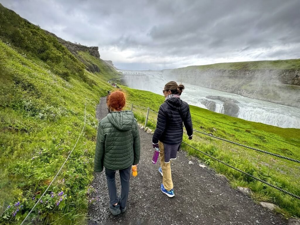 Viewers walk on a gravel trail beside a steaming geothermal area under cloudy skies. Lush green grass lines the path near the active landscape.