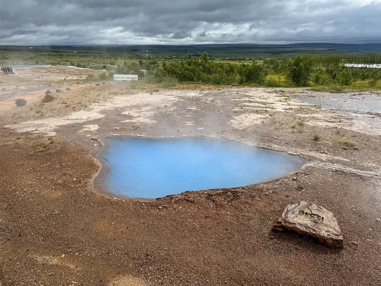 Geysir A vibrant blue hot spring set against a rugged geothermal landscape under an overcast sky.