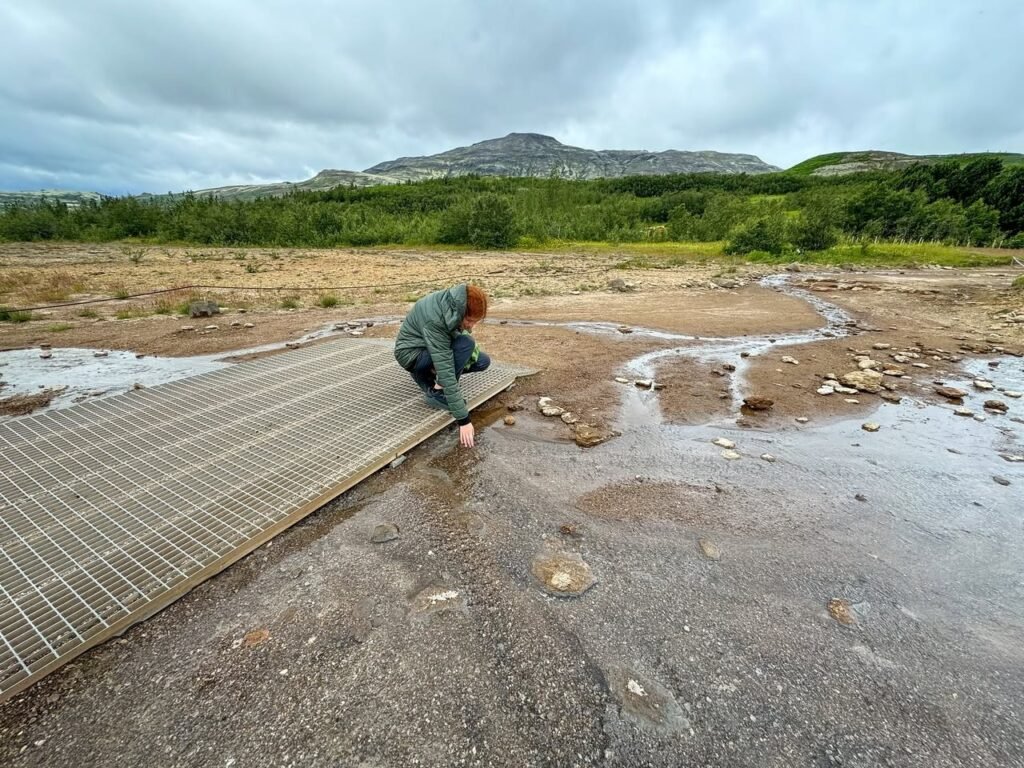 A person crouching by a metal ramp in a muddy creek bed with distant hills.