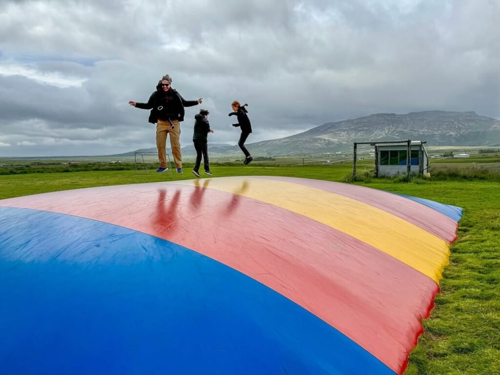 People jumping on a large, colorful, rainbow-striped inflatable airbeam in a grassy field with hills in the background under a cloudy sky.