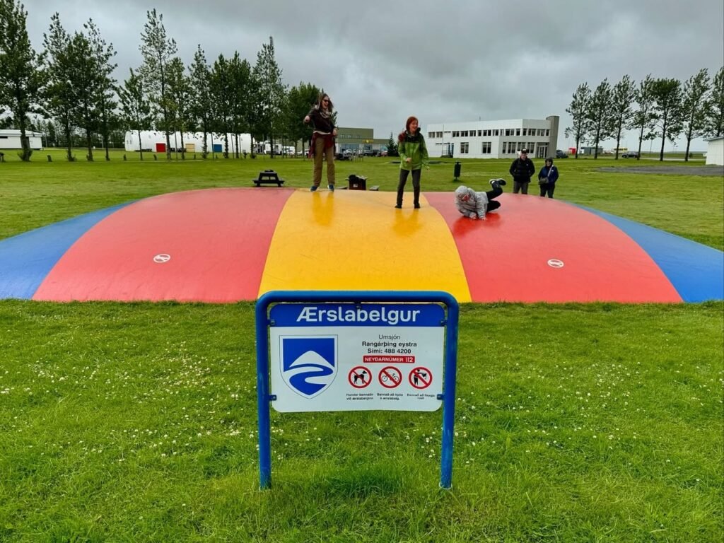 A vibrant, multi-colored inflatable structure set in a green park. A sign with Icelandic text and symbols stands prominently in the foreground.