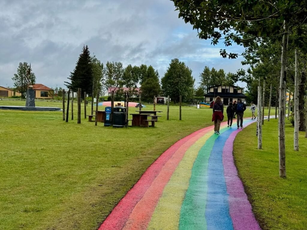 A vibrant rainbow-painted path winds through a grassy park, flanked by tall trees and a cloudy sky. A few people stroll along the colorful trail.