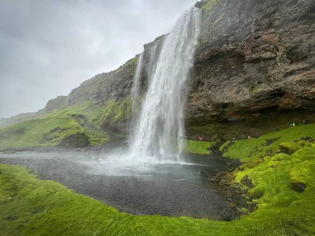 A tall waterfall cascades down a mossy cliff face into a dark pool surrounded by lush vegetation. Close-up views highlight the rugged texture of the surrounding rocks and vibrant green moss.