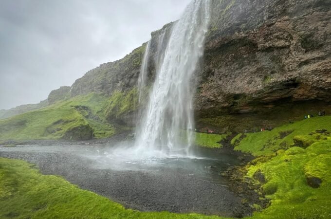 A tall waterfall cascades down a mossy cliff face into a dark pool surrounded by lush vegetation. Close-up views highlight the rugged texture of the surrounding rocks and vibrant green moss.
