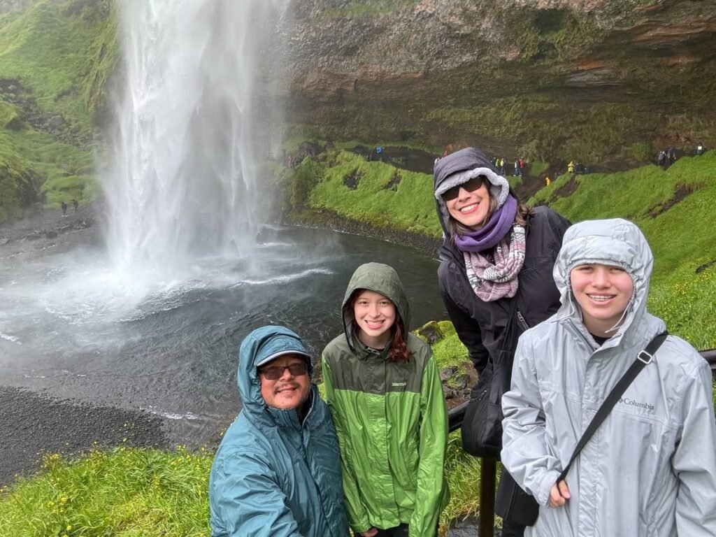 A group poses in rain jackets in front of a waterfall surrounded by lush greenery. A close-up highlights one traveler smiling in a hat and scarf.