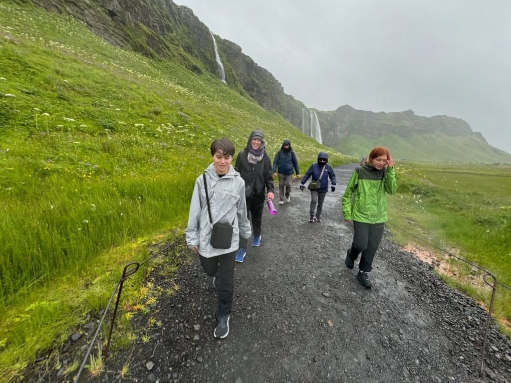 Hikers and a cyclist on a gravel path in a green landscape with distant mountains and waterfalls.