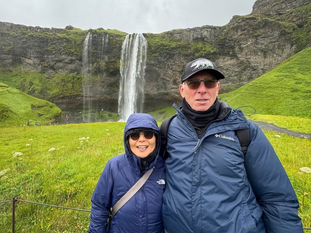 Visitors in rain gear stand before a tall waterfall with green grass and rocky cliffs.
