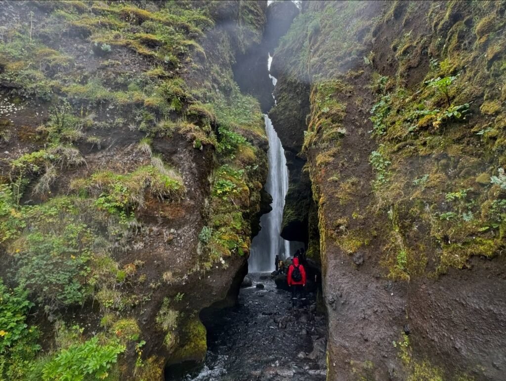 A person in a red jacket standing in the narrow fissure behind a hidden waterfall, surrounded by lush green moss-covered rocks.