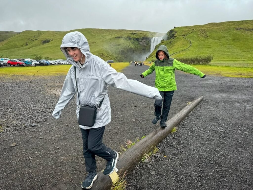 Two travelers in rain jackets walking on a log with a waterfall and green hills in the background.