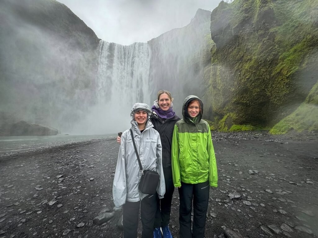 Travelers in rain jackets standing before a massive, mist-filled waterfall. This photo captures a moment of our world travel in a remote location, likely Iceland.