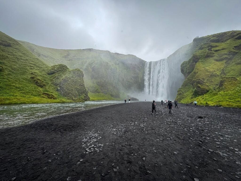 Lush green cliffs rise from a misty valley with a cascading waterfall visible in the distance. A dark gravel path leads through the serene natural landscape.