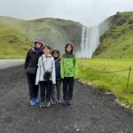 Puffins Travelers posing on a gravel path in front of a tall waterfall amidst green rolling hills on an overcast day.