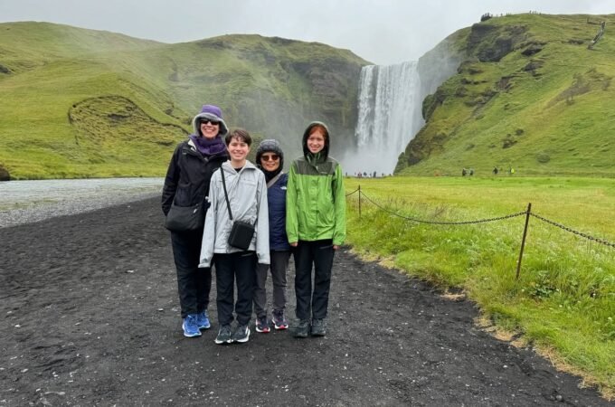 Travelers posing on a gravel path in front of a tall waterfall amidst green rolling hills on an overcast day.