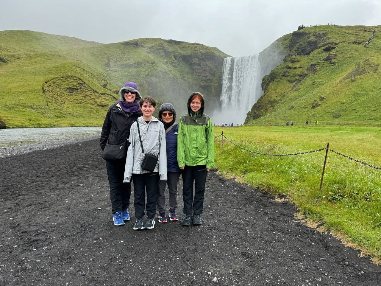 Skógafoss Travelers posing on a gravel path in front of a tall waterfall amidst green rolling hills on an overcast day.