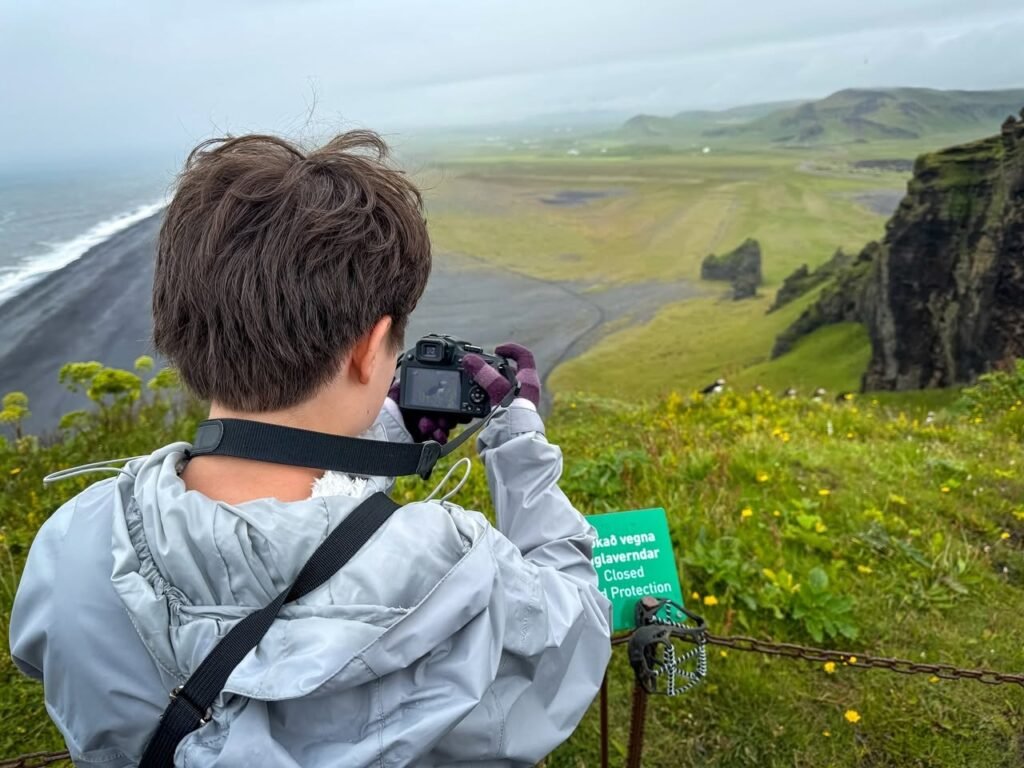 A photographer in a rain jacket captures the lush coastal cliffs overlooking the dark beach and ocean waves.