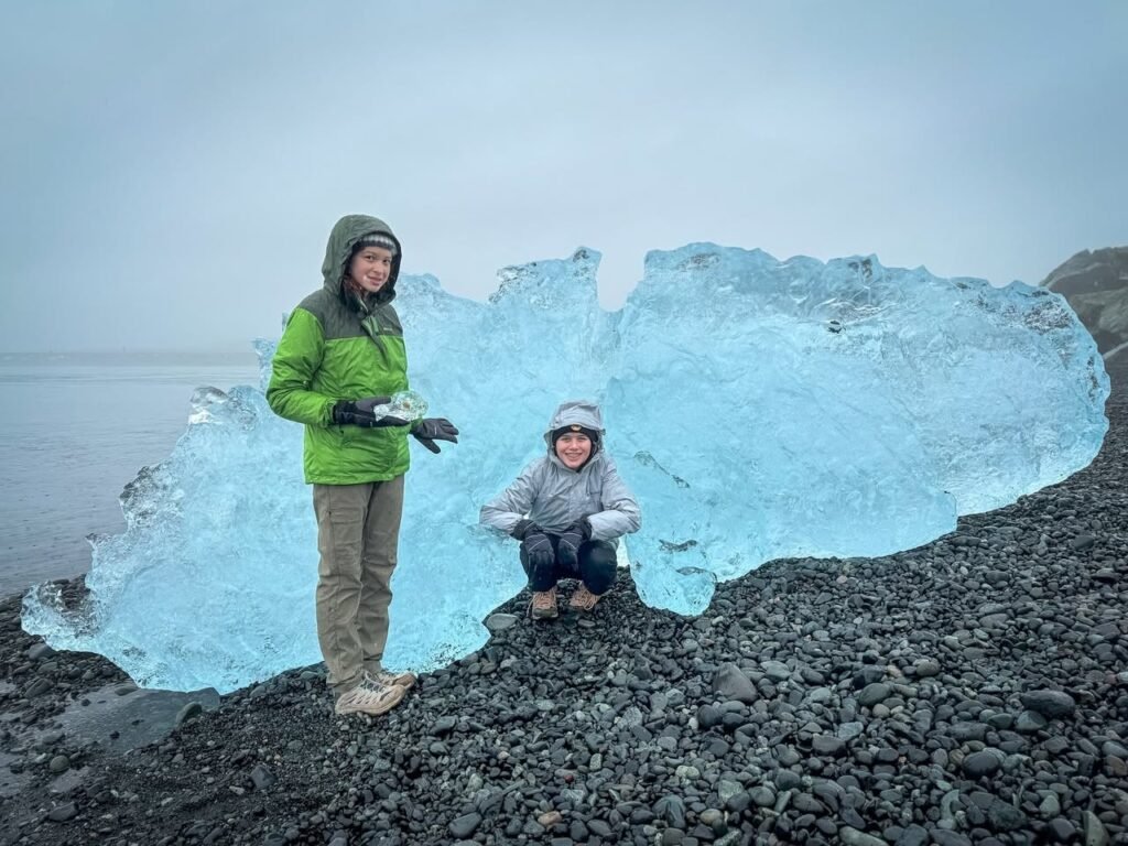 Two people posing with a large, glowing blue ice formation on a pebble beach under a cloudy sky.