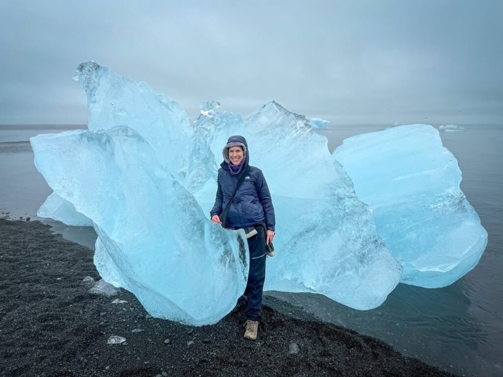 A traveler admires a stunning blue ice formation on a black volcanic beach.