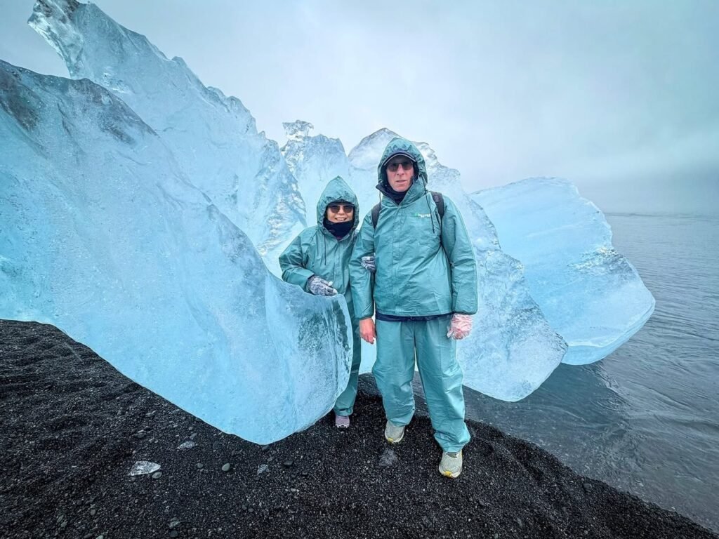 Two travelers in teal gear stand before a vibrant blue ice formation on a black sand beach. A companion photo shows a close-up view of mountainous ice textures.