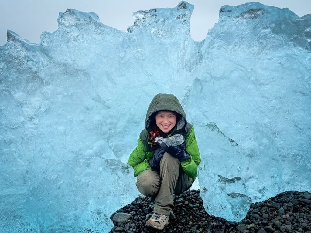 A young person in a green hooded coat sits against glowing blue ice. The colorful ice forms a natural backdrop for the portrait.