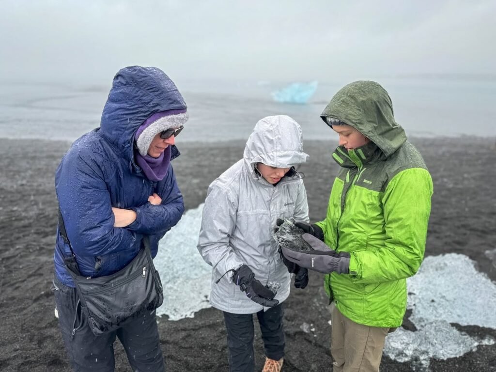 A group wearing bright rain gear stands near ice on a dark, misty beach. Heavy jacket hoods cover their faces as a glacier looms in the background.