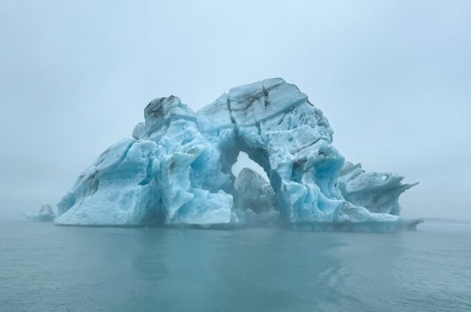 A large, sculpted blue iceberg featuring a natural arch floating in a misty arctic sea. A stunning polar scene captured during a remote travel expedition.
