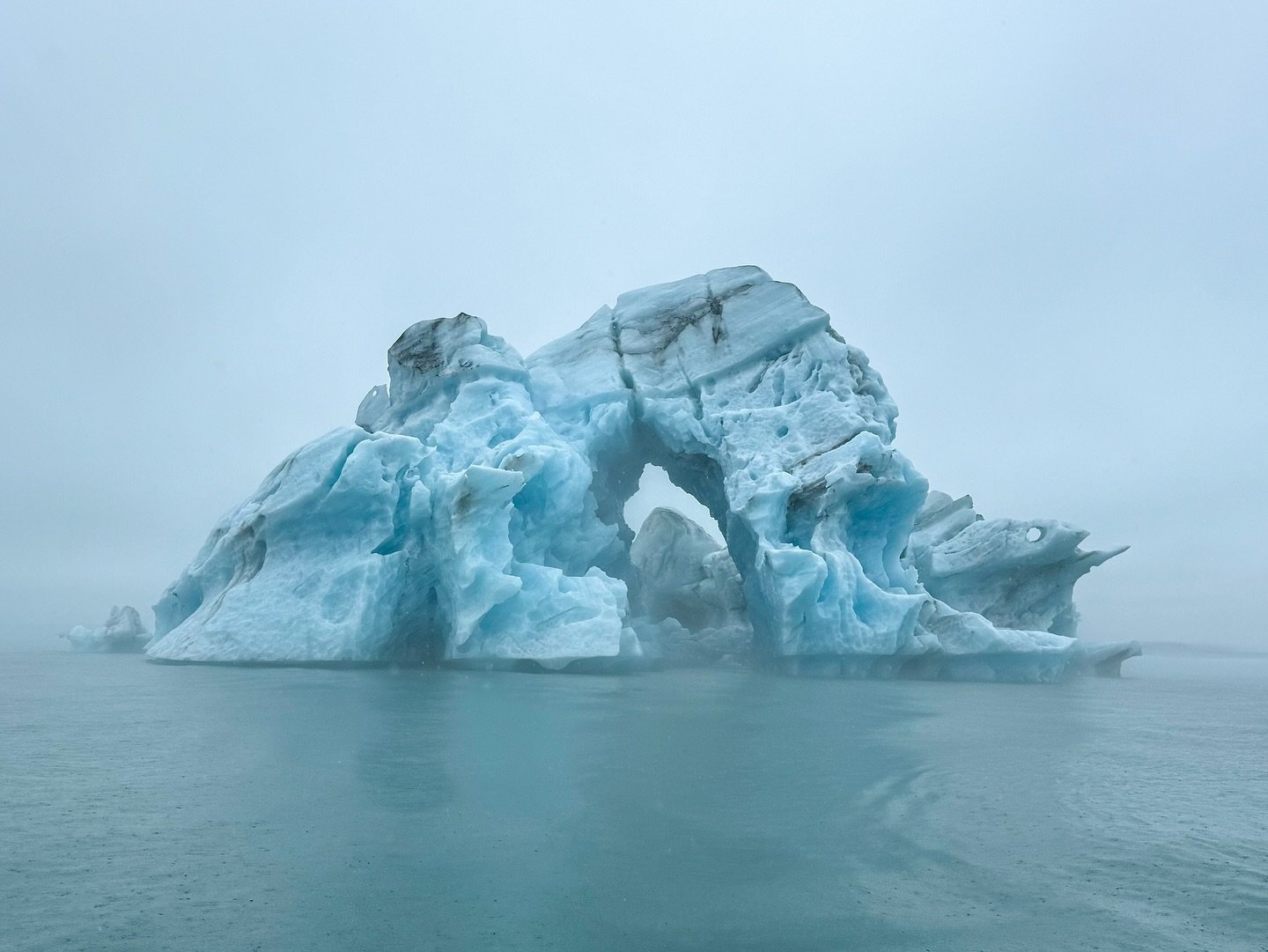 A large, sculpted blue iceberg featuring a natural arch floating in a misty arctic sea. A stunning polar scene captured during a remote travel expedition.