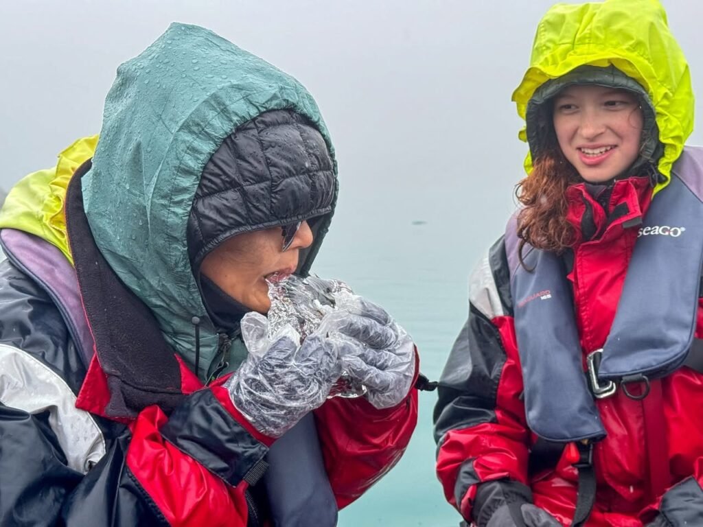 A person in a green hood and red jacket holds a container of water ice chips. Another individual wears a yellow hood and smiles against a misty background.