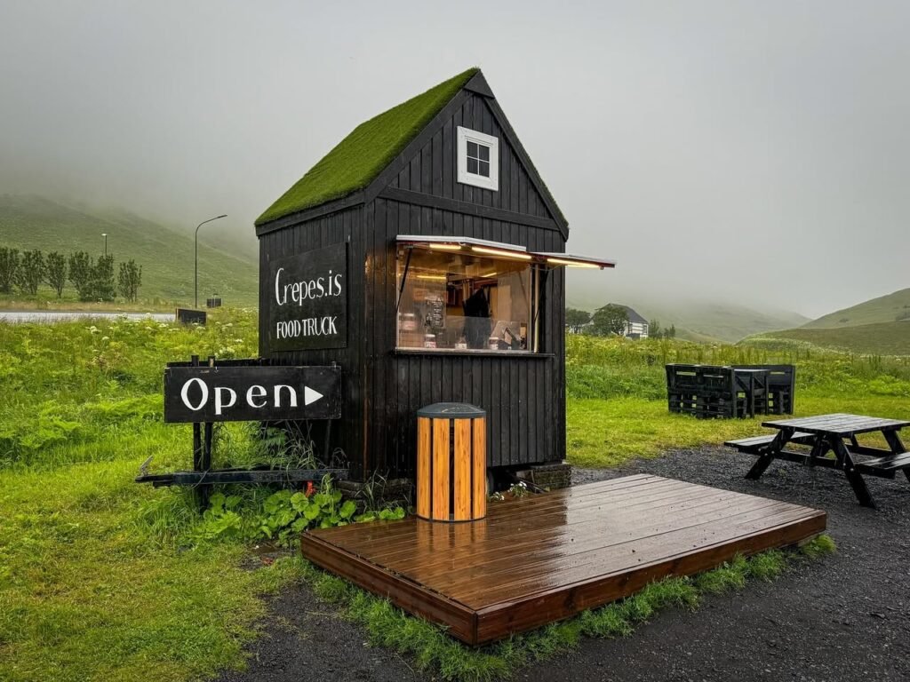 A charming Nordic cafe with a grass roof, featuring an open side service window surrounded by misty mountains and lush greenery.
