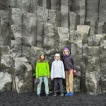 Three visitors stand in front of the basalt columns at the Giant's Causeway in Northern Ireland. The scene captures a family moment at a famous geological landmark.