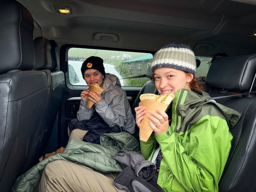 Two travelers wearing winter hats and jackets enjoy snacks from the backseat of a car. They share a smiling moment on a road trip.