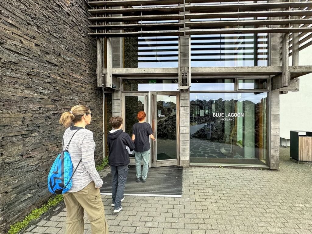 People entering a modern building with a textured slate wall and glass entrance. The architecture features horizontal wooden beams and large windows.