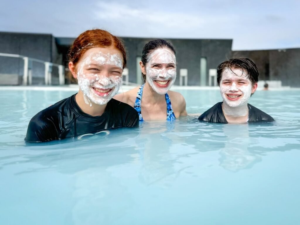 Three people swimming with white face masks in an infinity pool. One person is smiling with cream on their face.