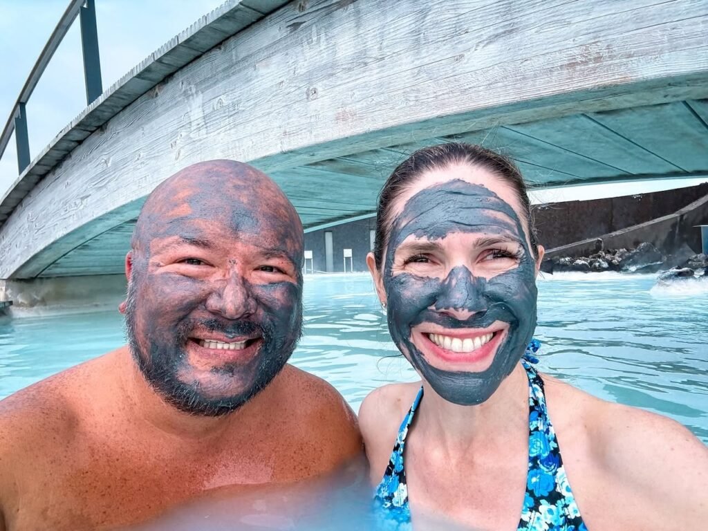 A smiling couple wearing dark mud face masks poses for a selfie under a bridge. The clear water and structural arches frame the couple's travel experience.