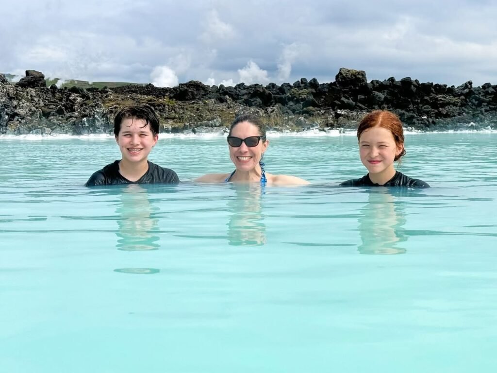 Three people swimming in calm, turquoise ocean waters near a rocky shoreline.
