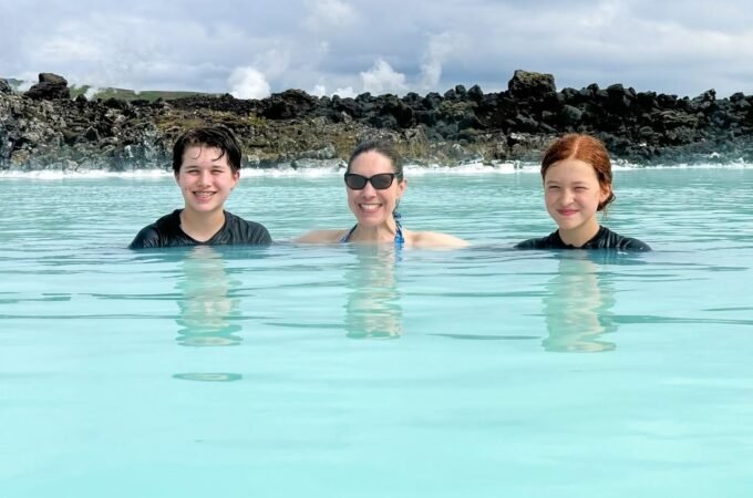 Three people swimming in calm, turquoise ocean waters near a rocky shoreline.