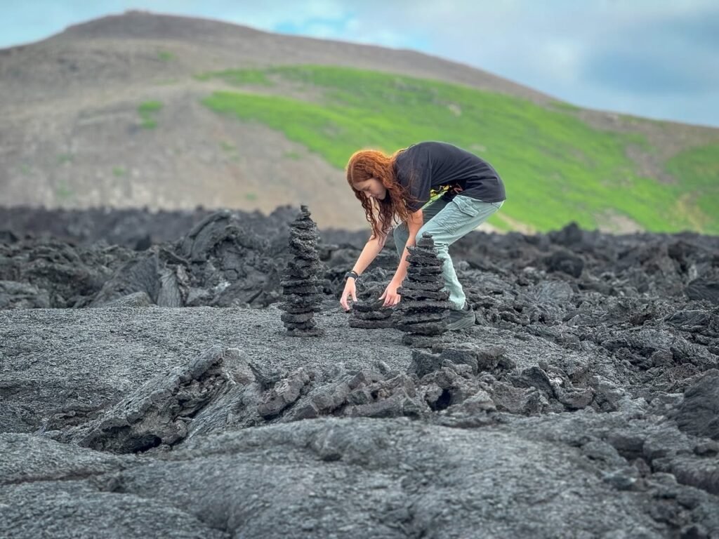 A traveler stacks small stones on a rugged lava rock field. Green slopes rise in the background beneath dramatic skies.